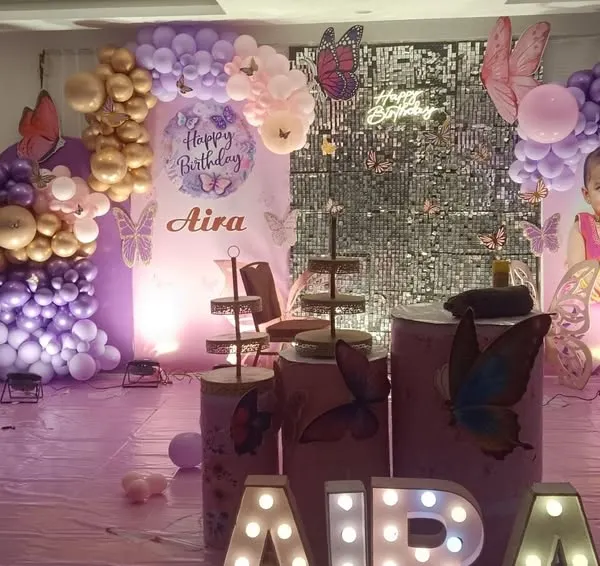 A beautiful butterfly theme for a first birthday, featuring shades of purple and pink. The decoration includes a silver shimmer wall, personalized backdrops with the child's photo, and marquee letters spelling her name.