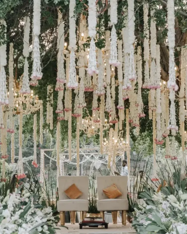 A beautiful mandap setup under a tree, featuring hanging garlands of white tuberose and a base of lush white flowers and greenery. The simple, elegant seating is perfect for an intimate and natural ceremony.