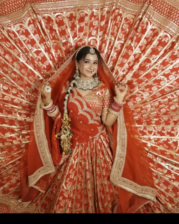 A full-length shot showcasing the grandeur of this traditional red bridal lehenga. The fanned-out dupatta with the 'Sada Saubhagyavati Bhava' border adds a classic, auspicious touch perfect for wedding photography.