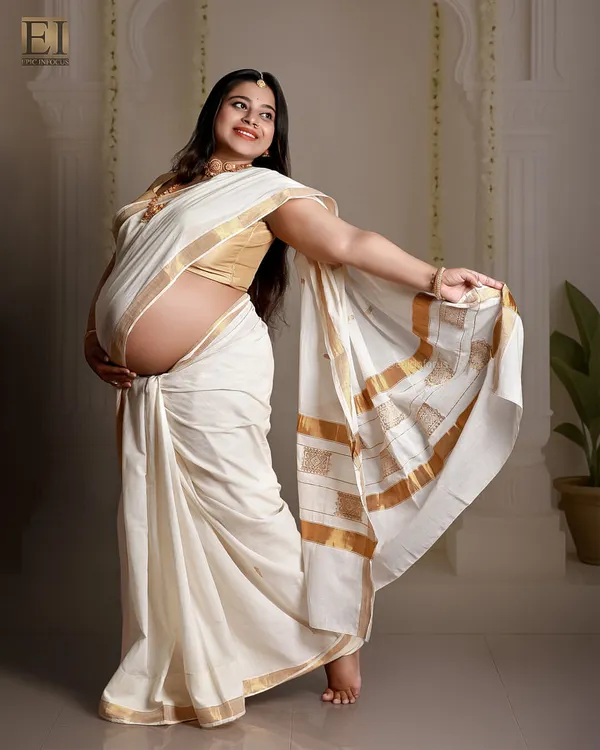 A joyful portrait of a mom-to-be showing off her baby bump in a classic white and gold Kerala saree.