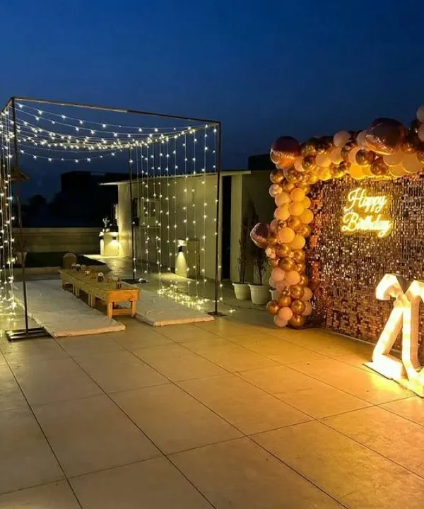 A stunning rooftop birthday celebration setup featuring a canopy of fairy lights, a shimmering gold sequin wall with a neon sign, and illuminated marquee numbers. This shows how I transform open spaces into magical event venues.