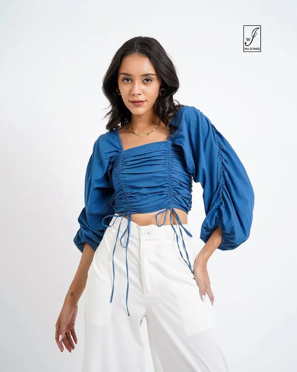 A crisp studio shot for a lookbook, featuring a model in a stylish blue ruched top and white trousers. The clean background ensures the clothing is the main focus.