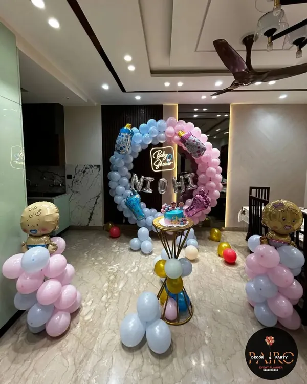 A full room view of a gender reveal or baby shower setup. The pink and blue balloon ring is the centerpiece, complemented by balloon columns topped with baby-shaped foil balloons.
