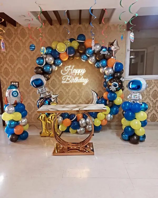 An out-of-this-world space-themed birthday party. This setup includes a balloon arch with planet and astronaut foil balloons, a neon sign, and a stylish cake table.