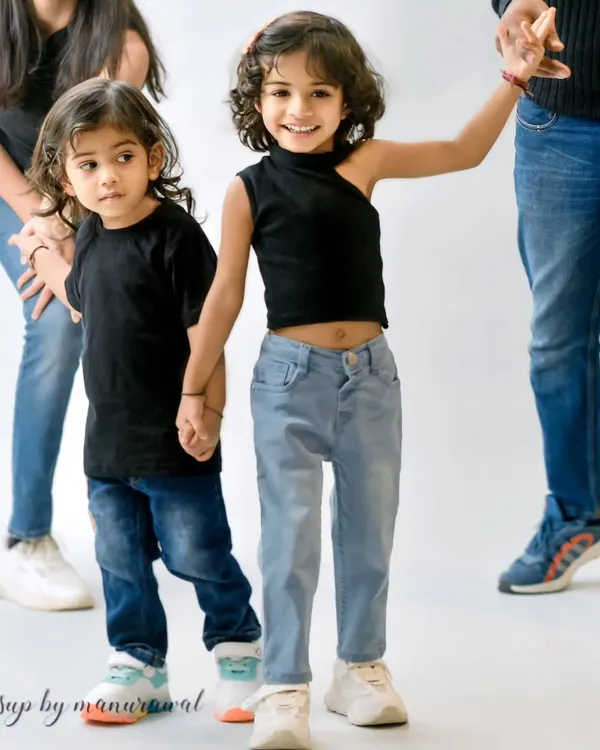 A candid and stylish shot of two young siblings holding hands in the studio, dressed in modern black tops and jeans, perfectly capturing their close bond.