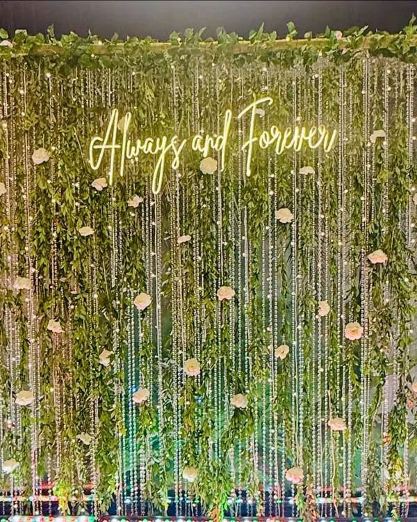 A romantic photo backdrop with a "Always and Forever" neon sign. The wall of cascading greenery, fairy lights, and scattered white roses creates an enchanting, garden-like effect.