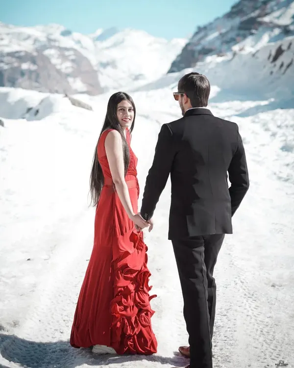 A classic romantic shot in the snows of Manali, with the bride in a striking red gown holding hands with her partner, creating a beautiful contrast against the white landscape.