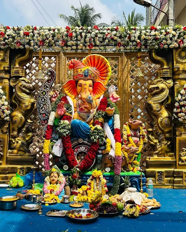 The final look of our grand Ganesh pandal, ready for worship. The large Ganesha idol is framed by ornate gold pillars and a lattice screen, with thick garlands of white chrysanthemums and red roses adding to the festive look.