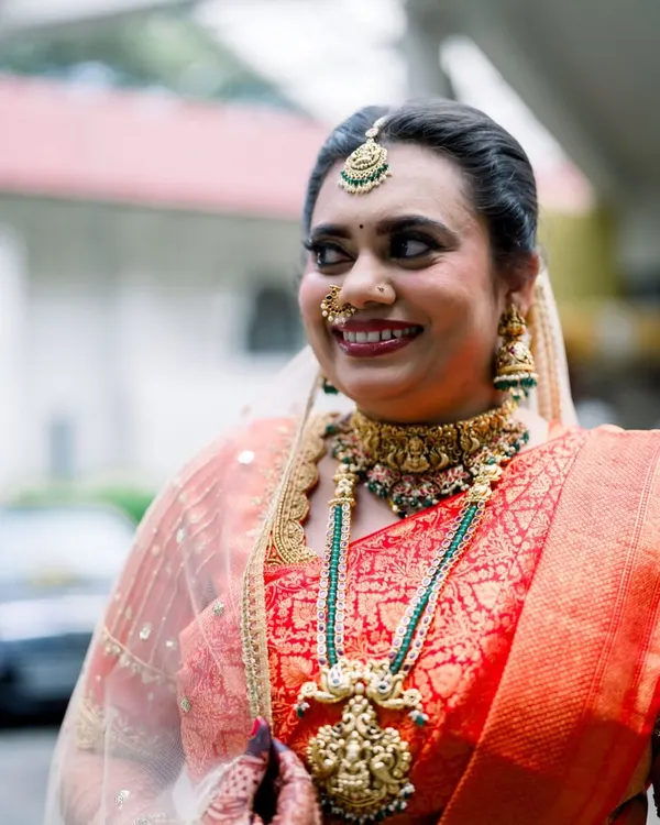 A radiant smile from our beautiful bride Suchetha. She chose a layered temple jewellery set with a statement choker and a longer haram featuring green bead accents, perfectly matching her stunning red bridal saree.