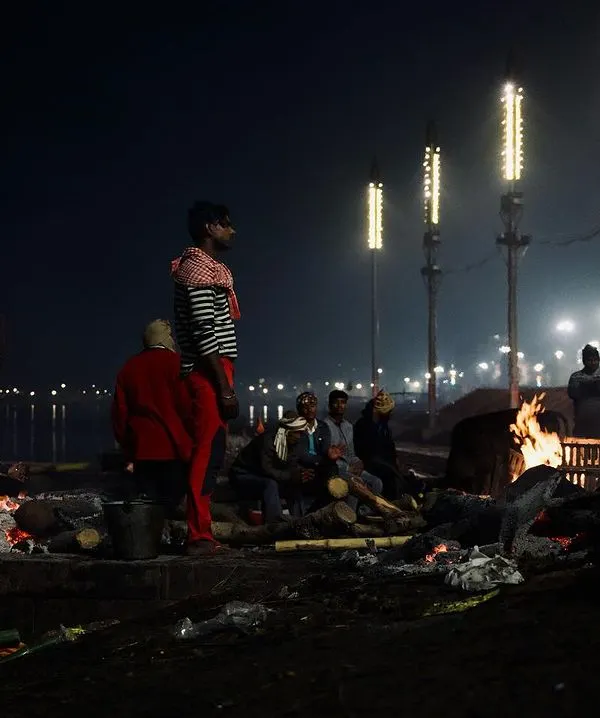 A wide night shot of Manikarnika Ghat, with multiple pyres burning against the dark waters of the Ganga. This image captures the scale of the ongoing cremation rituals that make this place a center of Hindu faith.
