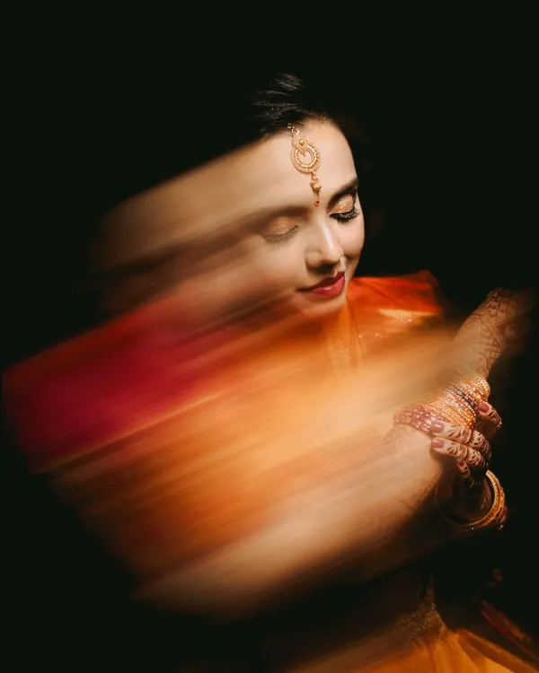An ethereal bridal portrait created with a slow shutter speed. The motion blur of her orange saree creates a beautiful, painterly effect, while her serene expression remains in focus, blending art with tradition.