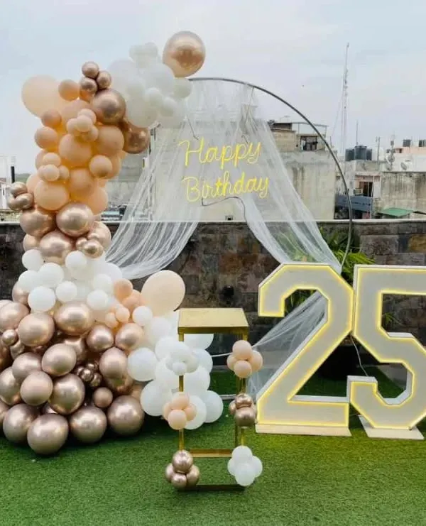 A 25th birthday celebration on a rooftop terrace. This setup features a semi-circular balloon arch in rose gold, peach, and white, a sheer canopy, a neon sign, and large marquee numbers.