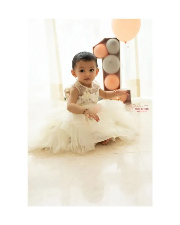A little princess in her beautiful white dress, celebrating her very first birthday. This minimalist studio setup with soft, natural light and pastel balloons puts all the focus on her sweet and curious expression.