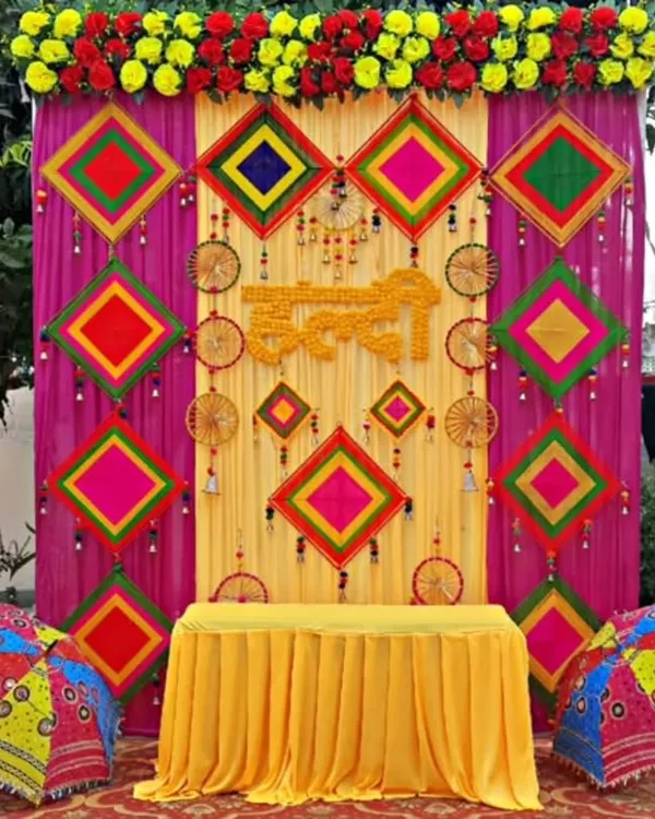 A truly grand Haldi backdrop featuring a vibrant mix of yellow and magenta drapes, decorated with colorful threadwork panels and dreamcatchers. The setup is framed with marigold garlands and Rajasthani-style umbrellas for a festive, traditional look.