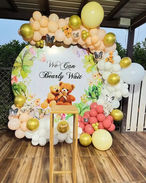 A charming teddy bear themed baby shower decoration with the message "We Can Bearly Wait". The setup features a custom circular backdrop framed by a balloon arch in pastel yellow, peach, and gold.