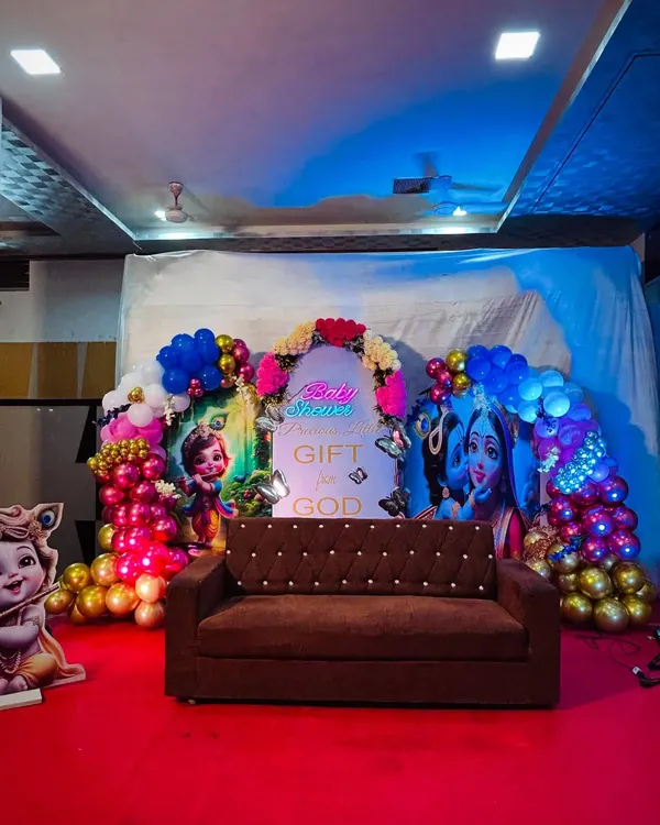 A full view of our grand Krishna themed baby shower stage. The setup includes two large character backdrops, a central panel with a blessing, a neon 'Baby Shower' sign, and a comfortable sofa for the parents to be, all set on a red carpeted stage.