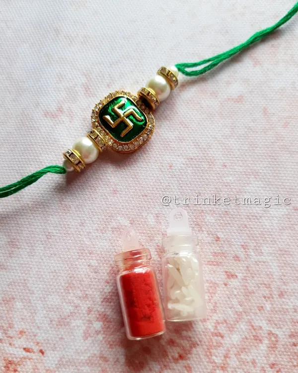 A simple and sacred Swastik Rakhi. This design features a green enamel Swastik symbol with pearl details on a traditional green thread, and it comes with a small bottle of Roli and Chawal.