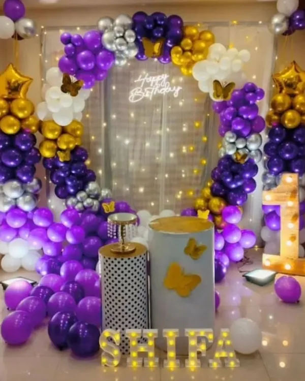 A rich purple, gold, and white theme with butterfly details. This setup includes a full balloon circle against a fairy light curtain, a light-up number '1', and marquee letters for the name 'SHILPA'.