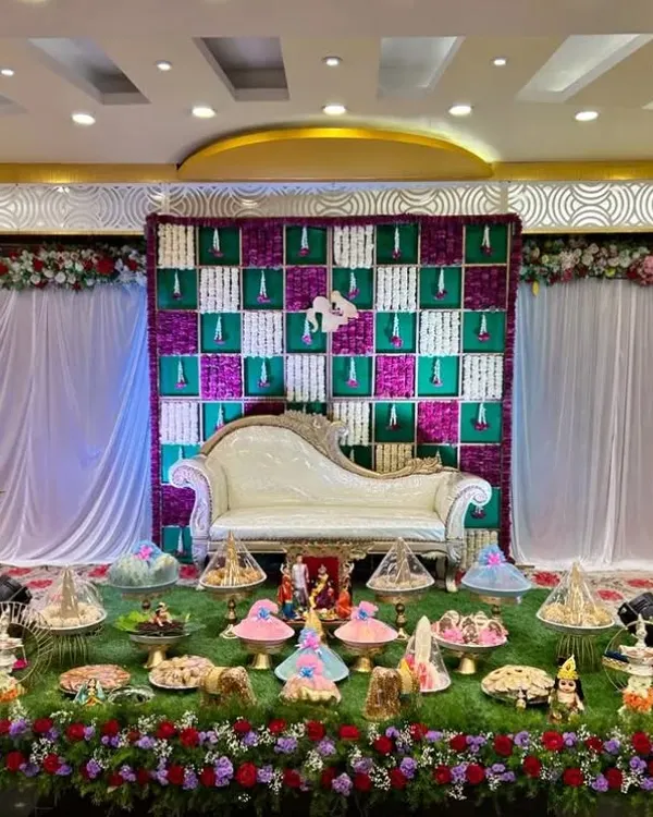 A full view of a grand Seemantham stage. The decor features a checkered floral backdrop in purple and white, a classic white chaise lounge, and an extensive display of decorated plates on an artificial grass carpet.