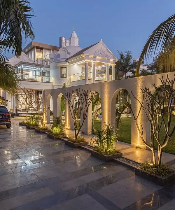 The 'Tropical Manor' at night, where landscape lighting transforms the space. The illuminated arches create a dramatic rhythm along the driveway, highlighting the carefully selected trees and framing views of the grand villa beyond.