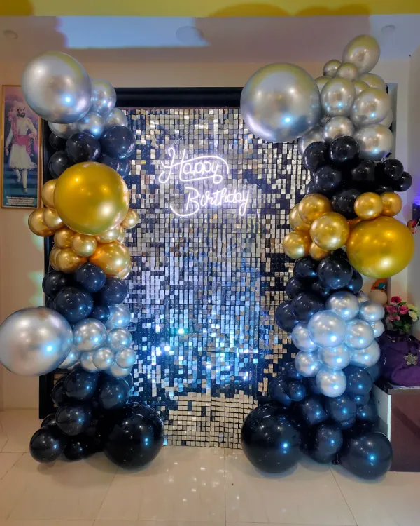 A close-up of our popular shimmer wall backdrop. This one in silver is framed by black, gold, and silver balloons and features a 'Happy Birthday' neon sign, creating a high-energy photo spot.
