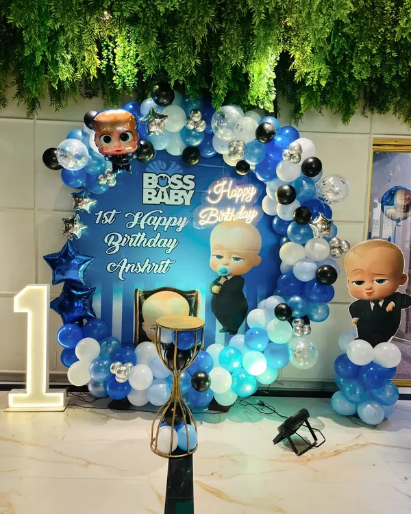 A "Boss Baby" themed first birthday party, featuring a custom backdrop with character cutouts, a blue, white, and silver balloon ring, and a light-up number '1'.