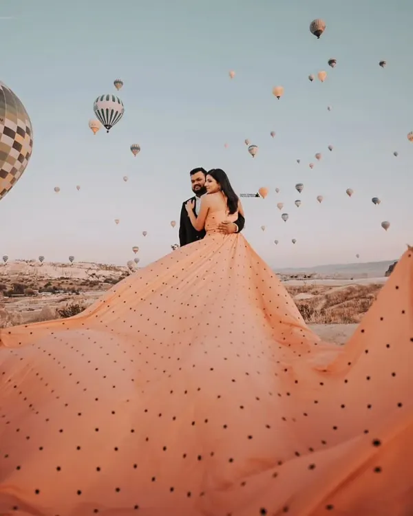 A breathtaking wide-angle shot showing the full expanse of the peach polka-dot gown's trail against the sky full of hot air balloons in Cappadocia. This is how you create a truly epic pre-wedding photo.