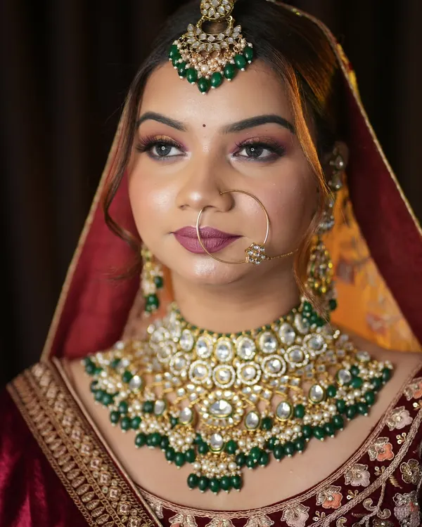 A classic North Indian bridal look featuring a deep red lehenga and stunning kundan jewelry with green accents. The makeup is focused on defined, smokey eyes and a bold lip to create a timeless and royal feel.