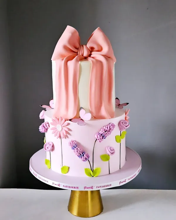 This elegant two-tiered cake is a perfect example of the popular "bow cake" trend. It features a tall top tier wrapped in a dramatic, handcrafted pink fondant bow, with the bottom tier decorated with delicate, piped wildflowers.