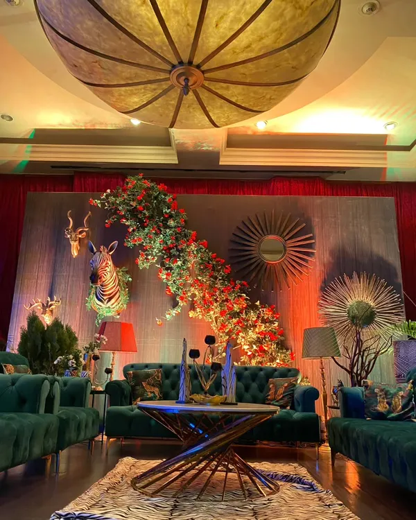 An enchanted forest Sangeet theme, featuring a lounge area with deep green velvet sofas, animal-print cushions, and a backdrop of faux animal heads and cascading florals.