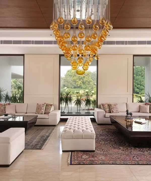 This expansive drawing room in a neo-classical farmhouse was designed to be filled with natural light. The large glazed openings connect the interior to the vast green landscape, while muted wall mouldings and a statement chandelier add a sense of timeless luxury.