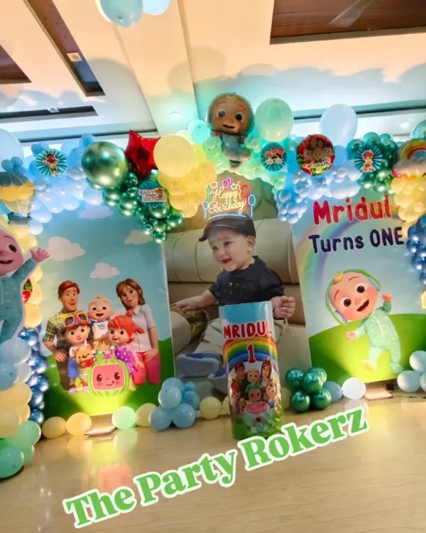 A complete Cocomelon-themed setup for a first birthday at home. It includes a balloon arch, a large backdrop with a photo of the birthday boy, character cutouts, and a light-up number.