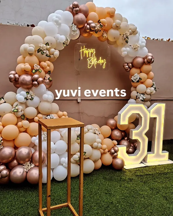 This is a chic outdoor setup for a 31st birthday, featuring a circular balloon arch in nude, white, and rose gold tones. The design is completed with a "Happy Birthday" neon sign and large light-up marquee numbers.
