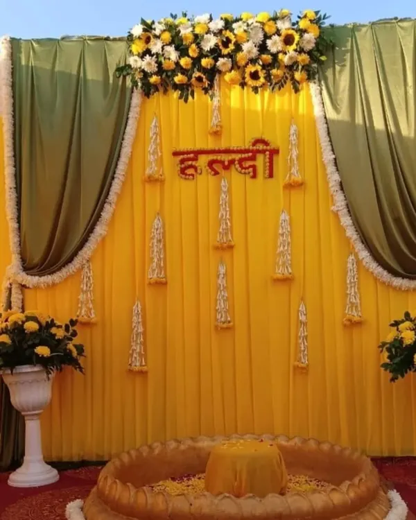 This is a classic Haldi ceremony backdrop I created for an outdoor event. The setup uses a bright yellow fabric base with contrasting green drapes, and the top is decorated with fresh sunflowers and white chrysanthemums for a vibrant look.