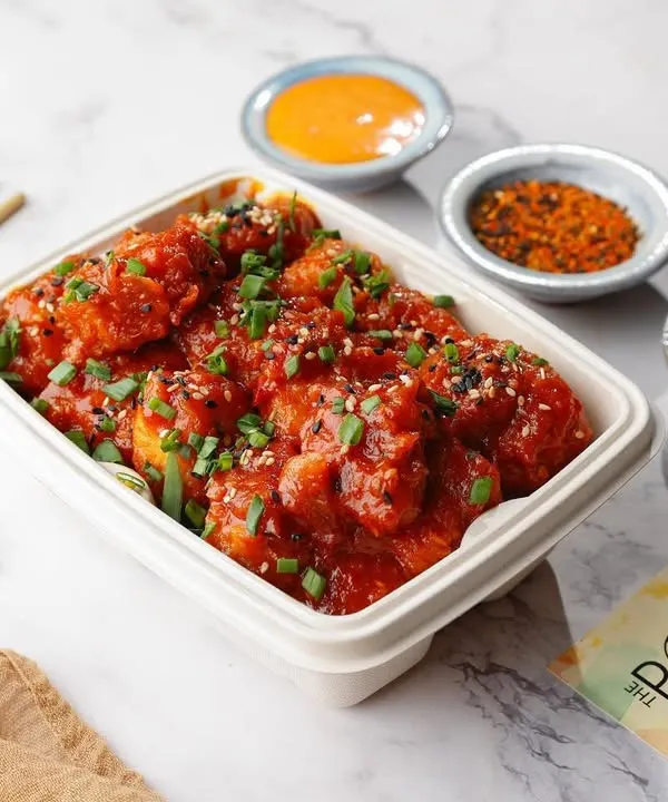 Saucy Korean fried chicken in a biodegradable takeaway container, styled with dipping sauces to look like a complete and tempting meal.