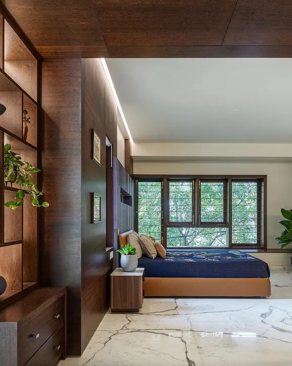 This bedroom in the Kanasu project is designed as a tranquil retreat. A custom wood-paneled wall incorporates a display niche and side table, while large windows frame the view of the trees outside.