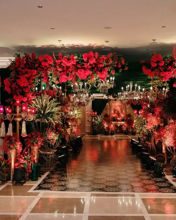 A grand entryway draped in a canopy of red blooms and illuminated by crystal chandeliers. This opulent passage sets a regal tone, welcoming guests into a celebration of timeless elegance.