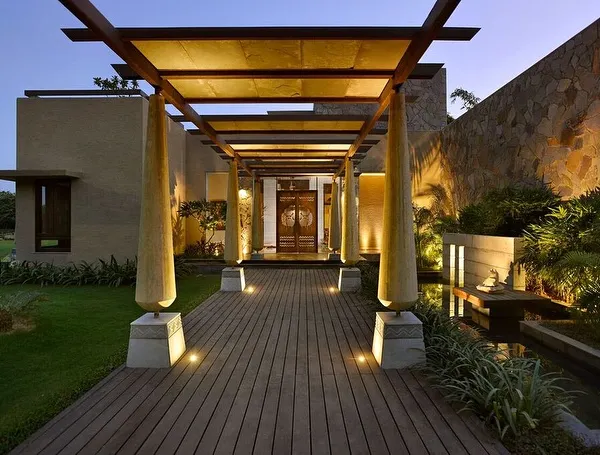 The entrance to C Farm in Chattarpur is marked by a wooden deck and a pergola supported by unique, cone-shaped columns. This walkway is flanked by a stone wall and a water channel, creating a multi-sensory journey into the home.
