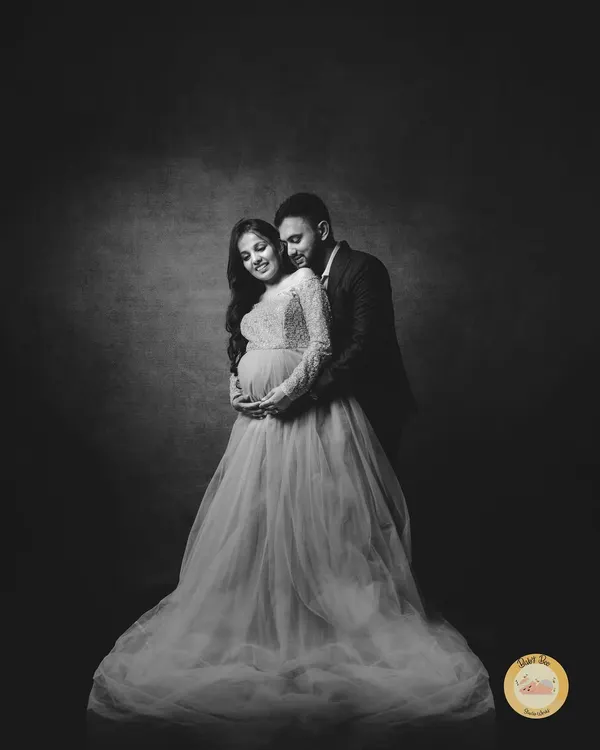 A dramatic and romantic black and white maternity portrait. The use of a dark, textured backdrop and a flowing gown creates a fine art feel, focusing on the couple's elegant pose and connection.