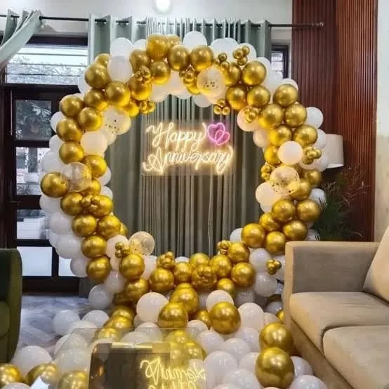 A stunning "Happy Anniversary" decoration for a home celebration. A large circular balloon arch made of gold and white balloons surrounds a neon sign, with more balloons filling the floor for a lavish look.