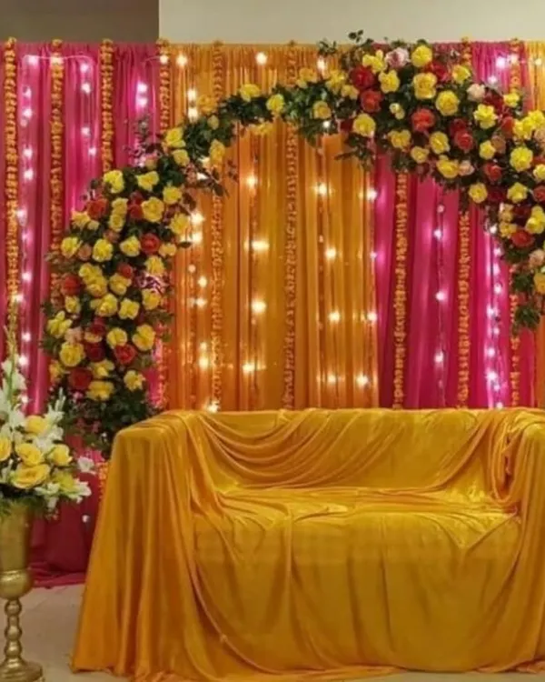 For pre-wedding functions like Haldi or Mehendi, I create vibrant backdrops that are perfect for photos. This setup features a beautiful arch of fresh yellow and red roses against rich pink and orange drapes, with fairy lights to add a warm glow.
