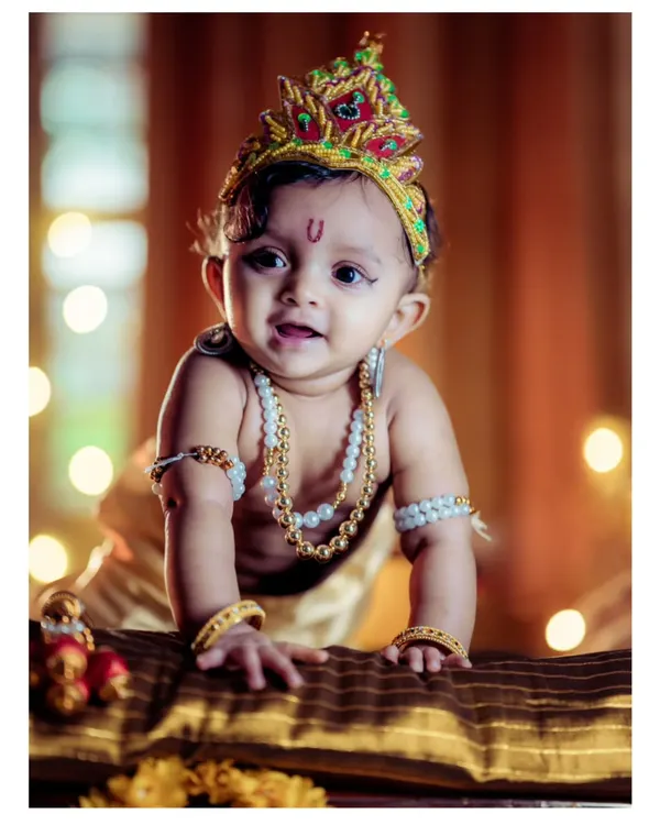 This portrait captures the bright, curious expression of a baby dressed as Lord Krishna. The warm, glowing lights in the background create a beautiful bokeh effect, and the detailed costume, from the peacock feather crown to the pearl necklaces, is arranged perfectly.