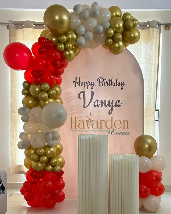A simple yet elegant birthday setup at home. The arched backdrop with a custom name and a red, gold, and white balloon garland creates a beautiful focal point.