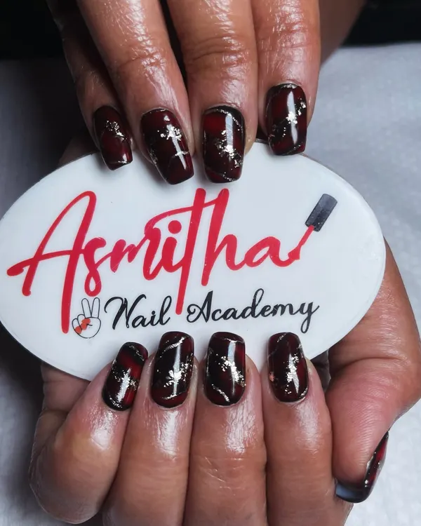 A detailed look at our red and black marble nail art, held against our Asmitha Nail Academy logo. The gold foil flakes add a touch of glamour to the design.