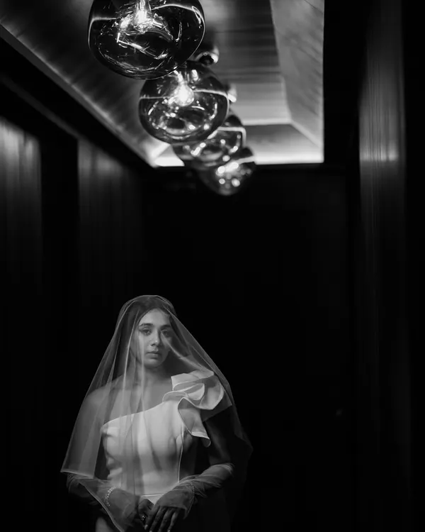 A bride veiled in shadow, illuminated by a row of vintage bulbs. This dramatic black and white portrait is a perfect example of how I use light and composition to create something truly artistic and memorable.