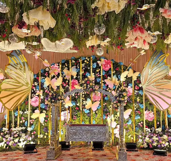 A full view of the enchanting butterfly-themed stage for a naming ceremony. The setup includes a silver cradle, a ceiling of hanging greenery and flowers, and large, shimmering butterfly wings.