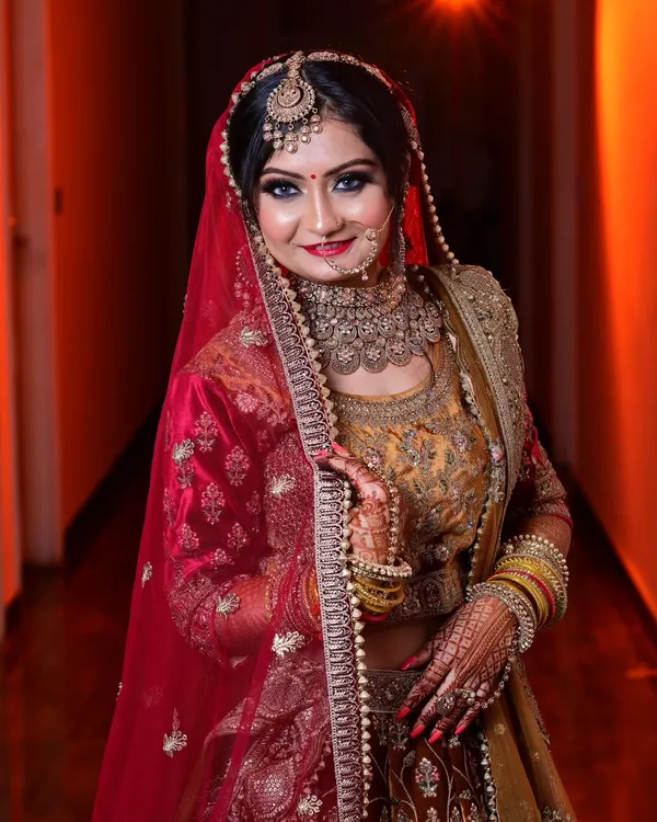 A classic bridal portrait showing the bride in her stunning red and gold lehenga. The warm lighting highlights her intricate jewelry and confident smile.