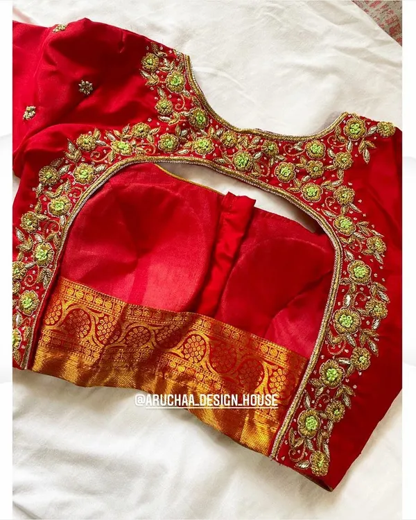 A stunning red bridal blouse with intricate floral embroidery in contrasting green and gold. The deep back cut is designed to be elegant and is perfect for pairing with a traditional silk saree.