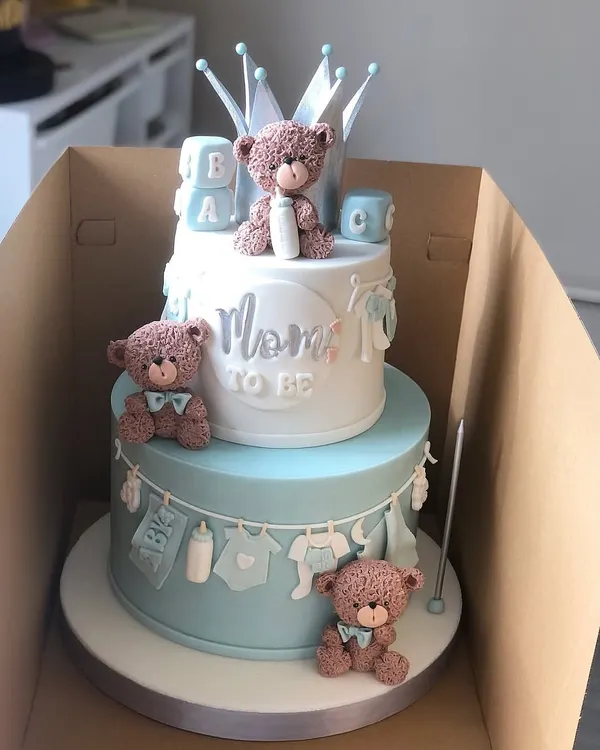 The cutest two-tiered "Mom to Be" cake for a baby shower. This design in pastel blue and white features adorable teddy bears, baby clothes, and a crown topper.