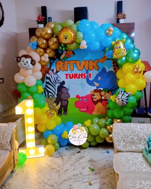 A fun jungle safari theme for a first birthday party at home. This setup includes a custom backdrop with cartoon animals, a colorful balloon arch, and a light-up number '1'.
